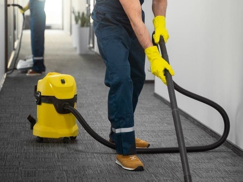 A cleaner vacuuming the carpet of a building corridor, a second cleaner working further down